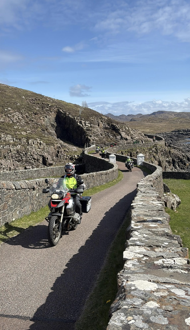 Riders travelling in formation along a narrow stone-lined scenic road.